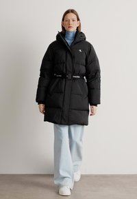 Giubbotto piumino nero con vita a cintura, dotato di un alto colletto e cappuccio. Tessuto liscio e design trapuntato. Abbinato a pantaloni svasati azzurri.
