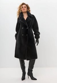 Schwarzer Kunstpelz-Mantel mit doppelter Knopfleiste, breitem Kragen und zwei großen Knöpfen. Getragen mit langen schwarzen Lederhandschuhen und kniefreien Stiefeln.