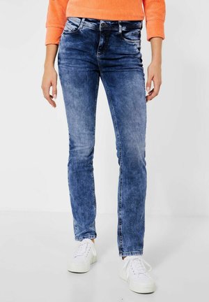 Jeans de mezclilla azules ajustados con un lavado desgastado, cinco bolsillos y un cierre de botón, combinados con zapatillas blancas sobre un fondo neutral.