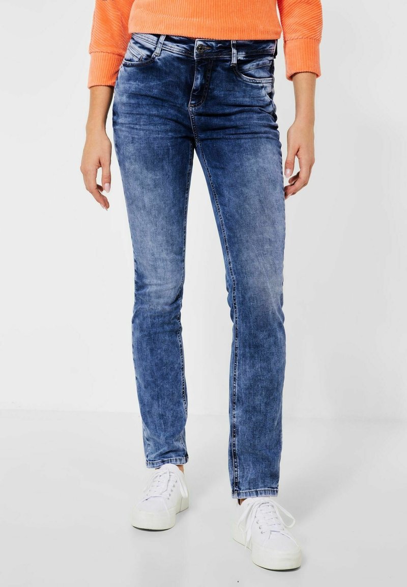 Slim-fit blå denimjeans med en blekt tvätt, fem fickor och knäppning, kombinerat med vita sneakers på en neutral bakgrund.