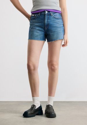 Άτομο που φοράει μπλε τζιν σορτς, λευκές κάλτσες, μαύρα μοκασίνια με λογότυπο, γκρι μπλουζάκι και ορατή μοβ ζώνη μέσης.