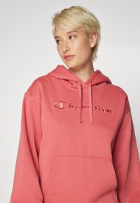 Felpa con cappuccio rosa realizzata in misto cotone; presenta una tasca a marsupio sul davanti e un logo "Champion" ricamato in una tonalità più scura.