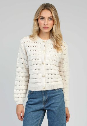 Cardigan en crème avec des motifs de dentelle verticaux et horizontaux, featuring un col rond et cinq grands boutons à l'avant.