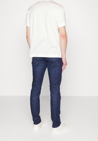 White cotton t-shirt, short sleeves, back view. Paired with dark blue denim jeans and white sneakers, displaying a slim fit design.