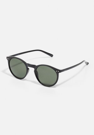 Lunettes de soleil rondes noires avec des verres teintés en vert, arborant un design épuré, des accents en métal et une finition mate sur les montures.