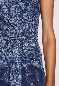 Dettaglio di un vestito floreale blu con cintura annodata in vita, che mostra motivi intricati e parte di un braccio nudo.