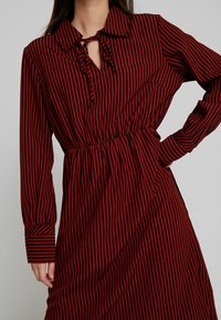 Femme portant une robe longue à manches longues rayée verticalement rouge et noire, avec un col et un détail de cravate au niveau de l'encolure, main sur la hanche.