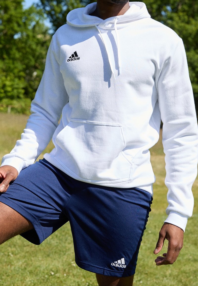 Bílá mikina s kapucí a přední kapsou, s černým logem, spárovaná s námořnickými sportovními kraťasy, které mají logo na lemu.