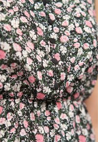 Robe à fleurs avec un fond noir, ornée de fleurs roses et blanches. Cinq boutons noirs décorent l'avant, avec une taille froncée.