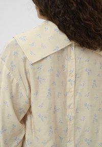 Svijetložuta košulja s velikim ovratnikom, koja sadrži suptilan plavi cvjetni uzorak. Dugmad su na leđima, a materijal je mekan i teksturiran.