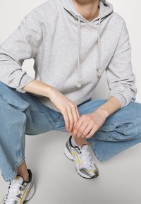 Felpa di cotone grigio chiaro con cordini e nappine; abbinata a jeans in denim blu pallido e larghi, e sneakers grigio-bianco con dettagli gialli.