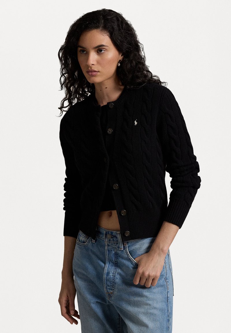 Cardigan negru tricotat cu model cu gât rotund, butoni pe față și detaliu mic cu logo. Mâneci și tiv cu model ribbed; purtat peste blugi albaștri deschiși.