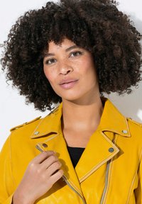 Chaqueta de moto de cuero amarilla con herrajes plateados, cierre de cremallera y epaulettes en los hombros, llevada sobre una camiseta negra. Cabello rizado y fondo neutro.