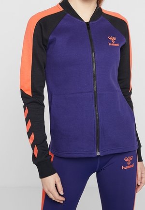 Frau trägt eine lila Jacke mit Reißverschluss und schwarzen sowie orangen Ärmeln, passende lila Leggings mit orangen Streifen und Hummel-Logo auf beiden Seiten.