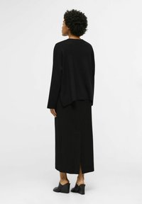 Maglione nero oversize con scollatura rotonda, abbinato a una lunga gonna nera con spacco posteriore; indossato con mule neri con tacco.