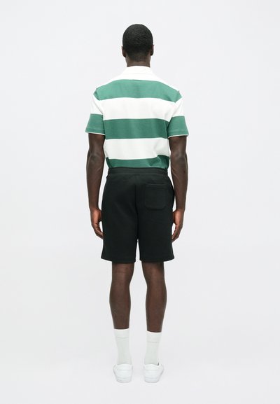 Zaļu un baltu svītrainu polo krekliņu apvieno ar melniem šortiem. Modelis valkā baltus sporta apavus un gaišas zeķes, redzams no aizmugures.