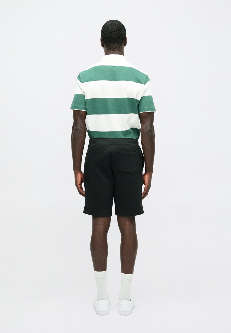 Maglietta polo a righe verde e bianca, abbinata a pantaloni corti neri. Il modello indossa sneakers bianche e calzini chiari, vista da dietro.