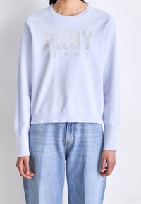 Ljusblå sweatshirt med ribbade ärmslut och rundad halsringning. Har en glänsande silverfärgad "DKNY JEANS"-logotyp över bröstet. Bärs tillsammans med jeans.