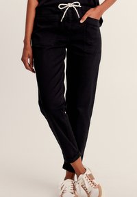 Schwarze Baumwoll-Jogginghose mit Kordelzugbund, zwei Fronttaschen und schmal zulaufenden Knöcheln. Getragen mit weißen und beigen Sneakers.