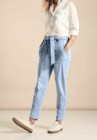 Pantalones ajustados de color azul claro con cintura con cinturón, dos bolsillos laterales y una textura suave, combinados con zapatillas blancas sobre un fondo neutro.