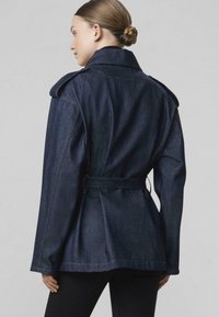 Žena so svetlými vlasmi zviazanými v uzle, oblečená v tmavomodrej rifľovej bunde s opaskom, zobrazená zozadu na jednoduchom svetlom pozadí.