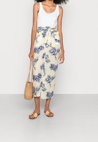 Blomsterindpakket nederdel i creme med blå botanisk tryk, med en sløjfe i taljen. Båret med en hvid tanktop og afslappede sandaler.