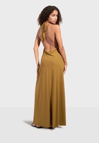 Femme aux cheveux bouclés portant une robe longue dos nu à col halter jaune moutarde et des sandales noires, se tenant devant un fond uni.