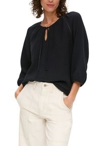 Czarna, luźna bluzka z okrągłym dekoltem i wiązaniem, z fakturowanej tkaniny, z rękawami o długości trzy czwarte, połączona z jasnymi spodniami.