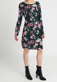 Bloemenjurk met lange mouwen, groene achtergrond, roze en witte bloemen, kniehoogte, met een zijstrikdetail. In combinatie met zwarte enkellaarsjes.