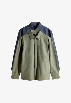 Zwei langärmlige Hemden zum Knöpfen, eines olivgrün vorne und eines dunkelblau mit Cordkragen dahinter, flach auf weißem Hintergrund ausgelegt.