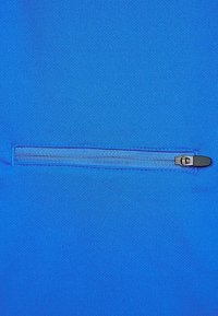 Sininen kangas, jossa on sileä pinta, sisältää vaakasuoran vetoketjullisen taskun. Vetoketjun vedin on musta ja metallinen, joka luo kontrastin siniselle materiaalille.