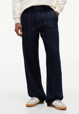 Άνδρας που φοράει φαρδιά σκούρα μπλε τζιν με μεγάλες μπροστινές τσέπες, συνδυασμένα με λευκά και μπεζ sneakers και κρεμ φούτερ.