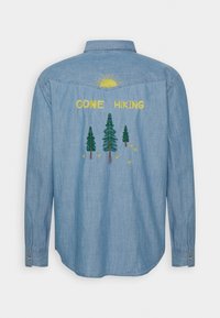 Light blue denim shirt featuring embroidered yellow sun and trees with "GONE HIKING" text. Long sleeves with button cuffs and curved hem.