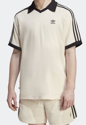 Homme portant un polo Adidas crème côtelé et un short assorti avec des rayures noires et des logos sur les manches et la jambe.