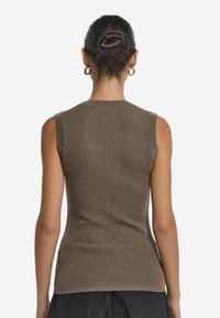 Kobieta z ciemnymi włosami związanymi w kok, nosząca złote okrągłe kolczyki, bezrękawowy brązowy top z prążkami i ciemne spodnie, pokazana z tyłu.