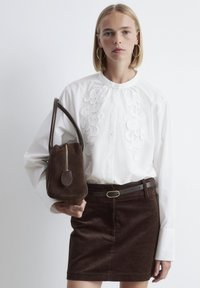 Woman with shoulder-length blonde hair wearing white embroidered blouse, brown corduroy skirt with black belt, and holding brown suede handbag.