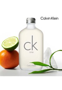 Bottiglia di vetro trasparente con tappo argentato, etichettata "ck one" e "Calvin Klein." Accompagnata da un lime verde, un'arancia, foglie verdi e ghiaccio.