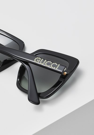 Melnas kaķa acs saulesbrilles ar gradienta lēcām, rotātas ar kristāla akcentiem, kas veido vārdu "Gucci" uz tempļa. Gluda plastmasas rāmja konstrukcija.