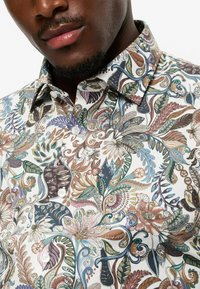 Patterned shirt featuring a white background with intricate floral and leaf designs in various colors, with a standard collar and button-up front.