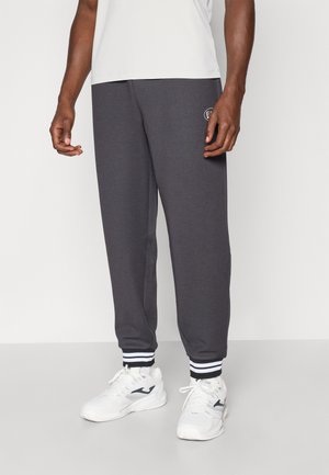 Pantalons de jogging en polaire grise avec des poignets rayés blancs et un patch logo. Portés avec des chaussures de sport blanches. Texture lisse et coupe décontractée.