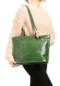 Borsa tote in pelle verde con una finitura liscia, forma rettangolare e due manici, tenuta da una persona che indossa un maglione lavorato a maglia beige e pantaloni neri.
