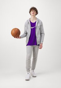 Haut de survêtement gris, débardeur violet avec logo Nike blanc, joggings gris et baskets blanches. Le modèle tient un ballon de basketball.