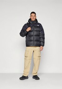 Μαύρο μονωμένο μπουφάν puffer με κουκούλα και φερμουάρ, με λογότυπο στο στήθος, σε συνδυασμό με μπεζ παντελόνι cargo και μαύρα παπούτσια.