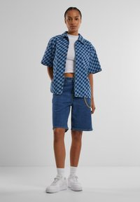 Blaues Karomuster-Hemd mit kurzen Ärmeln, weißes Croptop, blaue Denim-Bermudashorts mit Kettendetails und weiße Sneakers mit strukturierten Sohlen.