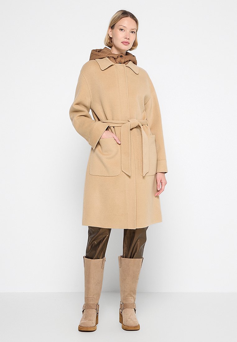 Abrigo de lana beige con cintura ceñida, grandes bolsillos y capucha forrada en marrón. Combinado con pantalones de imitación cuero marrones y botines beige.