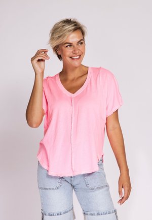 Smiling woman with short blonde hair wears loose pink V-neck shirt and light blue denim shorts, standing against plain background.