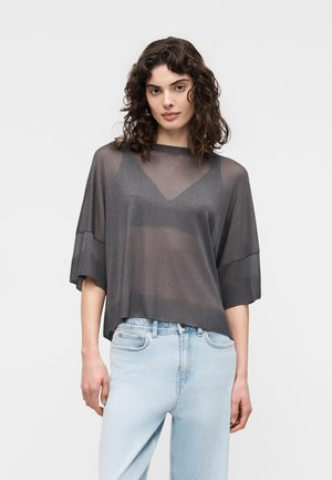 Donna con capelli ricci scuri che indossa una maglia grigia trasparente e larga sopra un bralette scuro e jeans azzurri, in piedi davanti a uno sfondo semplice.