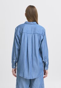 Camicia blu in chambray a maniche lunghe caratterizzata da strisce verticali bianche, colletto button-down e polsini bottone, con un design sul retro plissettato.