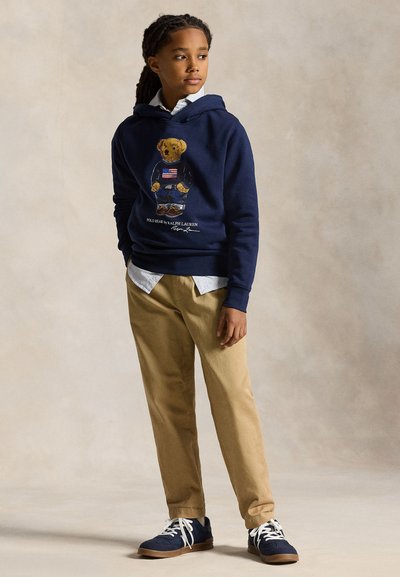 Poika, jolla on lettipäinen tukka, pukeutunut laivastonsiniseen huppariin, jossa on karhukuvitus, vaaleansiniseen paitaan, khakin värisiin housuihin ja laivastonsinisiin tennareihin, seisoo yksivärisen taustan edessä.