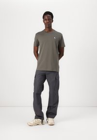 Tricou scurt de culoare verde măsliniu, cu un mic logo albastru, asortat cu pantaloni cargo gri care au buzunare laterale și o croială lejeră. Adidași bej.